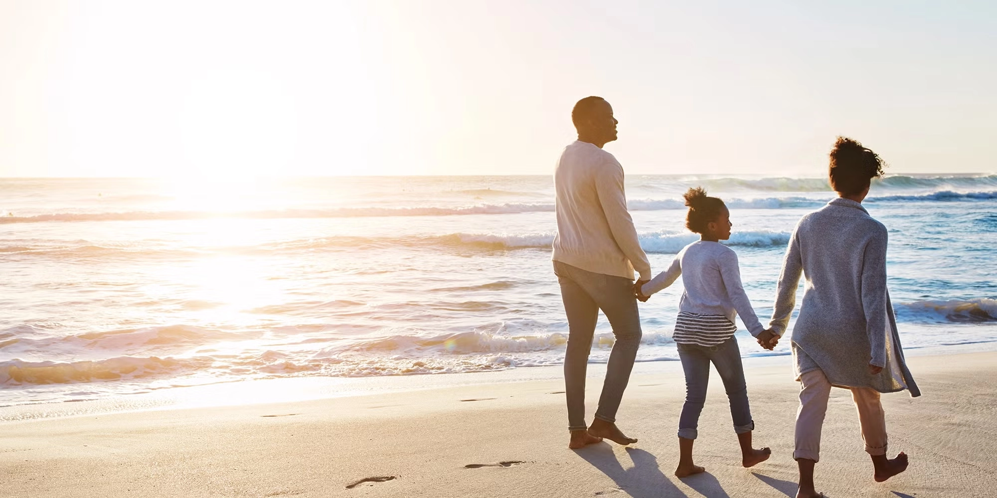 A family of three holds hands walking down the beach