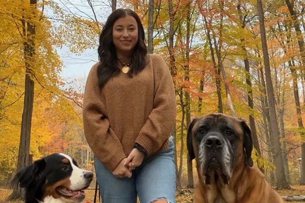 Taylor Hyma stands outside with her two dogs in a yellow autumn forest