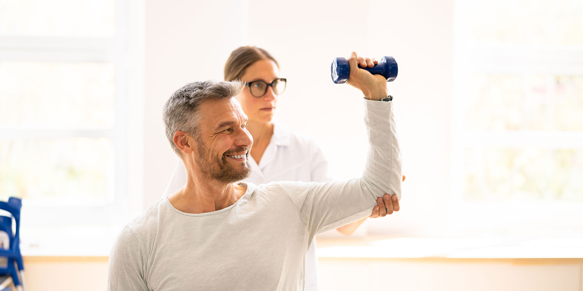 A smiling man raises his left arm by the elbow and holds a blue weight in his hand. Behind him, a female physical therapist guides him in his exercises