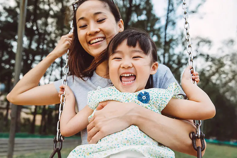 A mother holds her daughter as they swing at the playground while both of them laugh gleefully